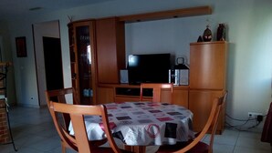 A dining area with a table covered by a patterned tablecloth, surrounded by wooden chairs with red cushions. A television is mounted on the wall above a wooden cabinet. - Close to center, comfort room in air-conditioned villa sauna, billiard, garden (Perpignan)