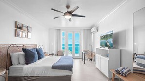 Un dormitori modern amb un llit, un ventilador de sostre, una televisió i un balcó amb vista al mar.