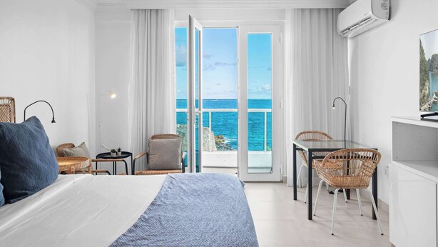 Habitació d'hotel amb un llit, escriptori, cadires de vimetla, una taula petita amb una planta i vista al mar a través de grans portes de vidre.