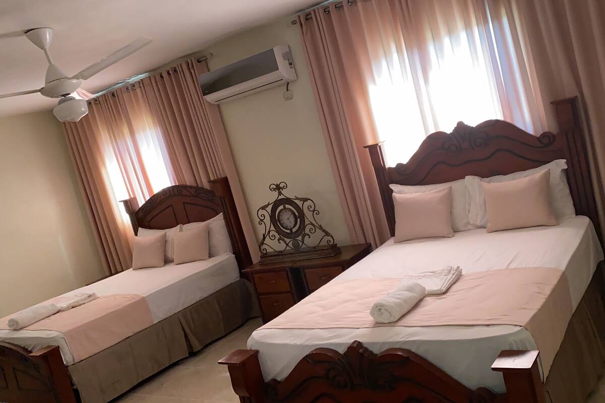 Habitación de hotel con dos camas individuales, ventilador de techo, equipo de aire acondicionado, mesita de noche con reloj y una planta en maceta.