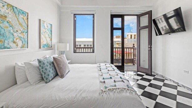 A bedroom with a bed, a TV, and a balcony with a view of buildings. - Mint Suite | 1BR Apt at Old San Juan (San Juan)