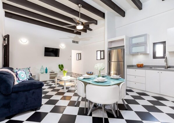 A modern kitchen with a black and white checkered floor, a dining table with chairs, a ceiling fan, and a TV mounted on the wall.
