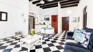 Una sala de estar moderna con piso a cuadros blanco y negro, un sofá oscuro con cojines estampados, una mesa de centro blanca con un jarrón de flores y un ventilador de techo.