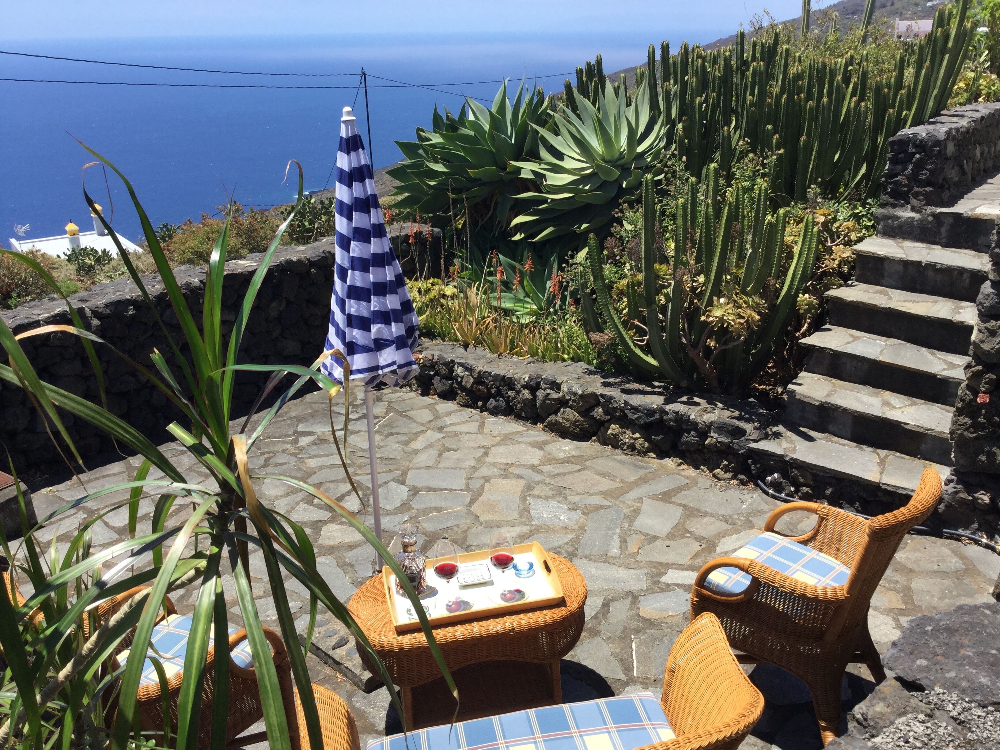 A patio with wicker furniture, a checkered umbrella, and a view of the sea.