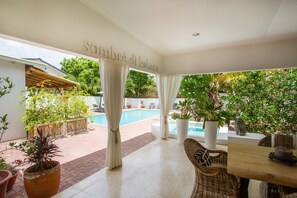 Un espacio al aire libre techado con piscina, muebles de mimbre, plantas en macetas y un letrero que dice "Sombré di Kabana".