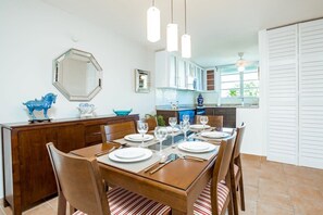 Een eetruimte met een houten tafel gedekt voor zes personen, omringd door stoelen. Achtergrond is een keuken met witte kasten en een raam.