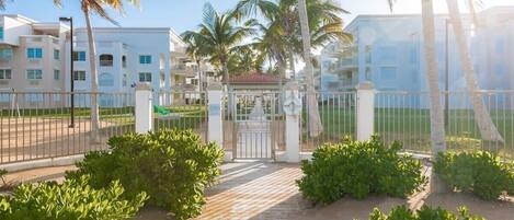 A gated entrance to a residential area with a wooden pathway leading to a gate, surrounded by palm trees and greenery.