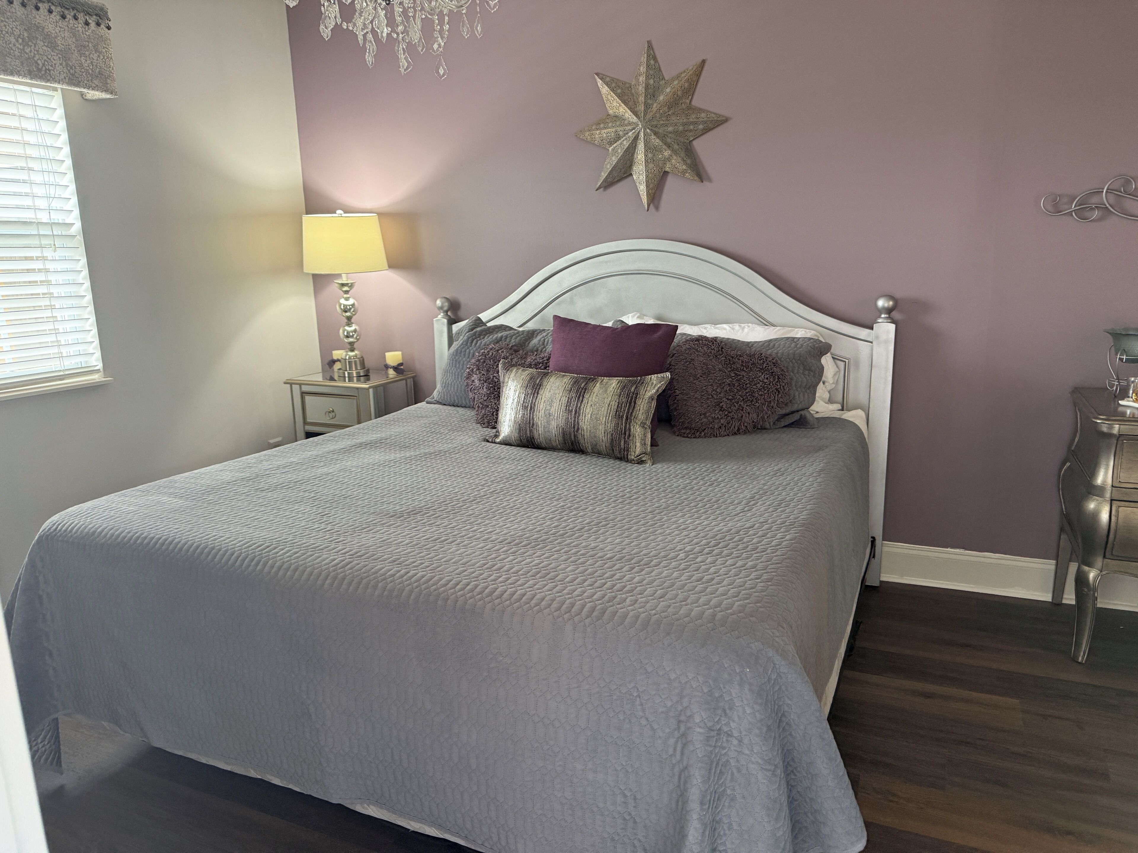 A neatly made bed with a grey quilt, a star decoration on the wall, and a lamp on a bedside table.