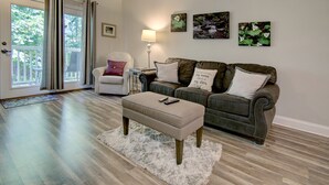 A living room with a beige ottoman, a brown leather sofa, a white armchair, and a lamp. There are three framed pictures on the wall. - INDOOR POOL! CLOSE TO DOLLYWOOD!! (Pigeon Forge)