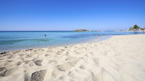 Een zandstrand met helderblauw water en een verafgelegen rotsformatie.