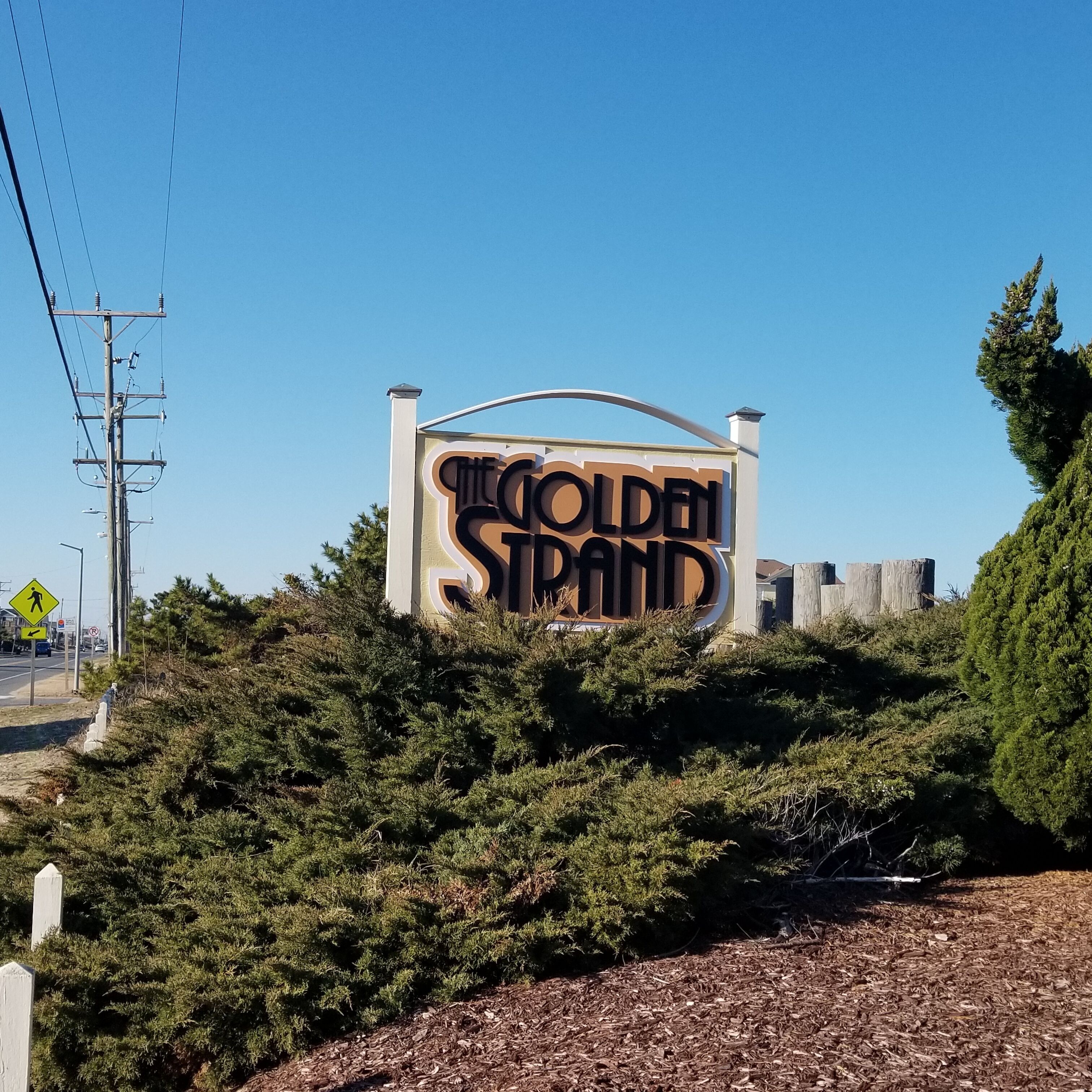 A sign for 'The Golden Strand' with a clear view of the surrounding area including a road, utility poles, and dense shrubbery.