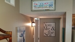 Un pasillo con una escena de playa enmarcada, un letrero en la pared que dice 'You Me & the Sea', y una silla de madera.