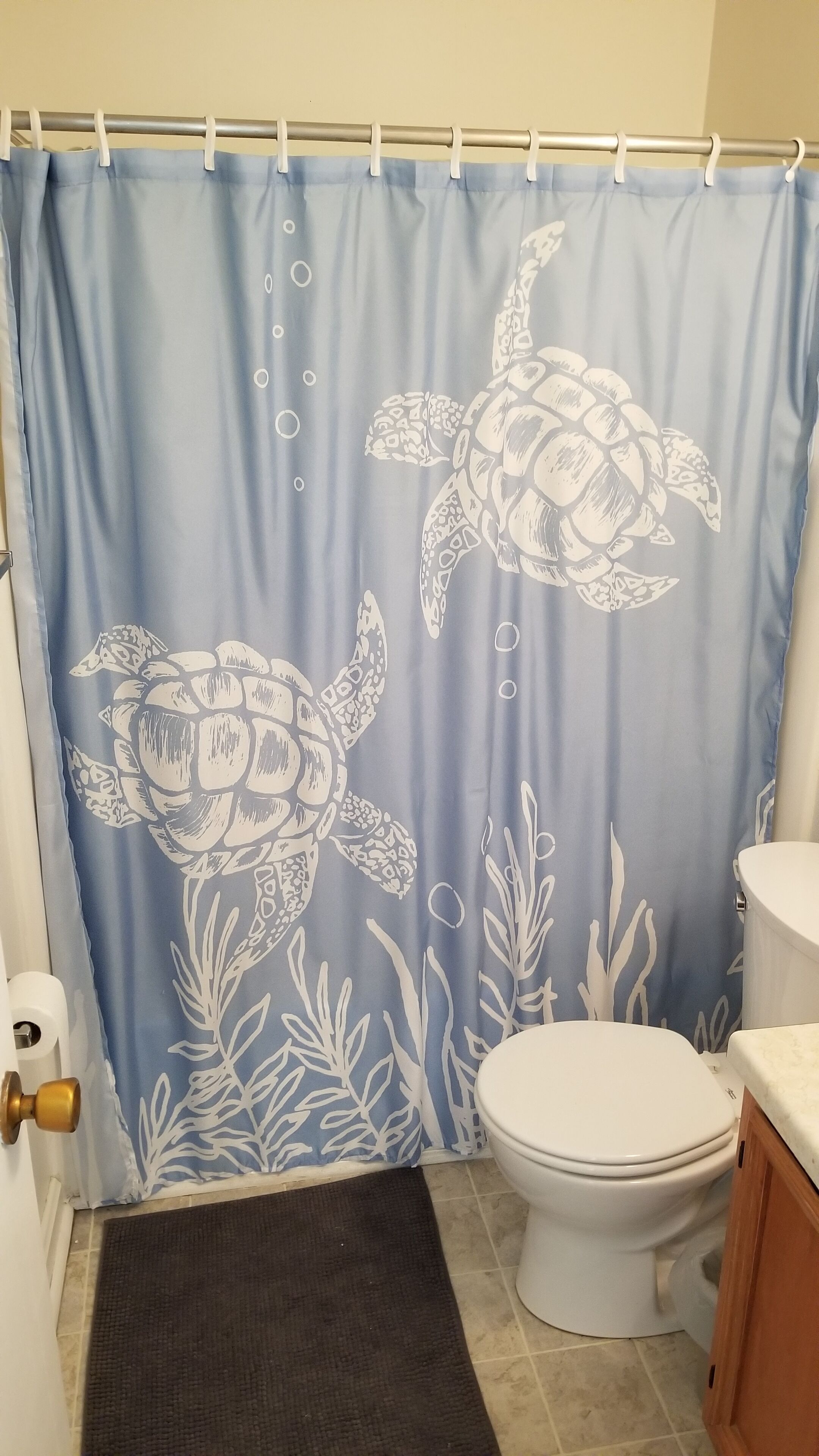 Une salle de bain avec un rideau de douche bleu orné de motifs de tortues de mer, des toilettes et un tapis marron.