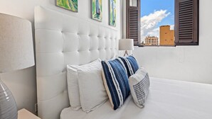 A hotel room with a white tufted headboard, a large window with wooden shutters, and three colorful paintings on the wall. - Azure Suite | 1 BR Apt at Old San Juan (San Juan)