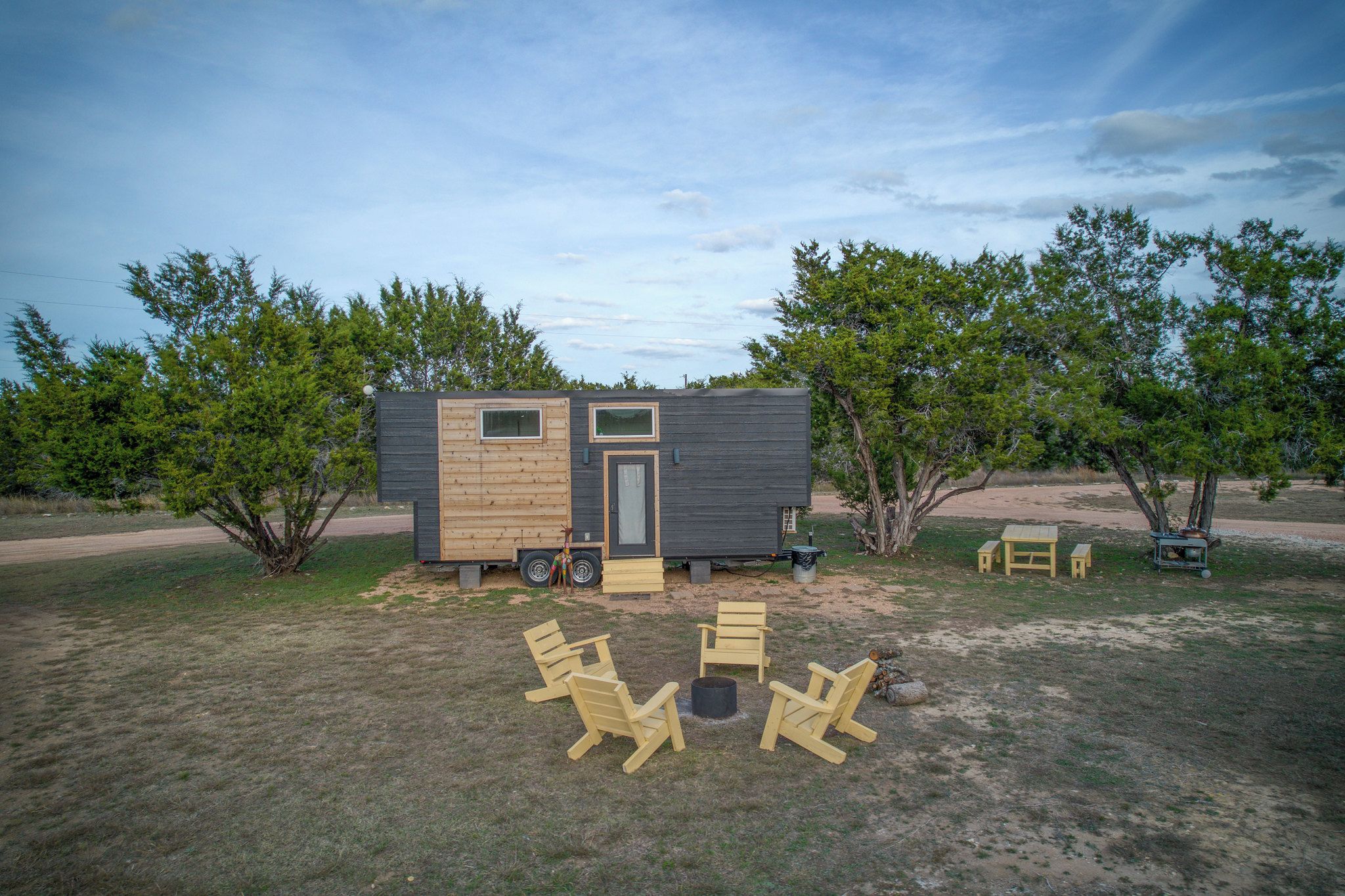Uma cabana pequena com porta e janelas, cercada por árvores e área de estar externa.