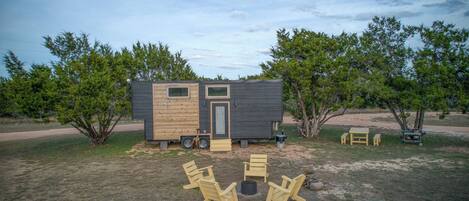 Uma cabana pequena com porta e janelas, cercada por árvores e área de estar externa.