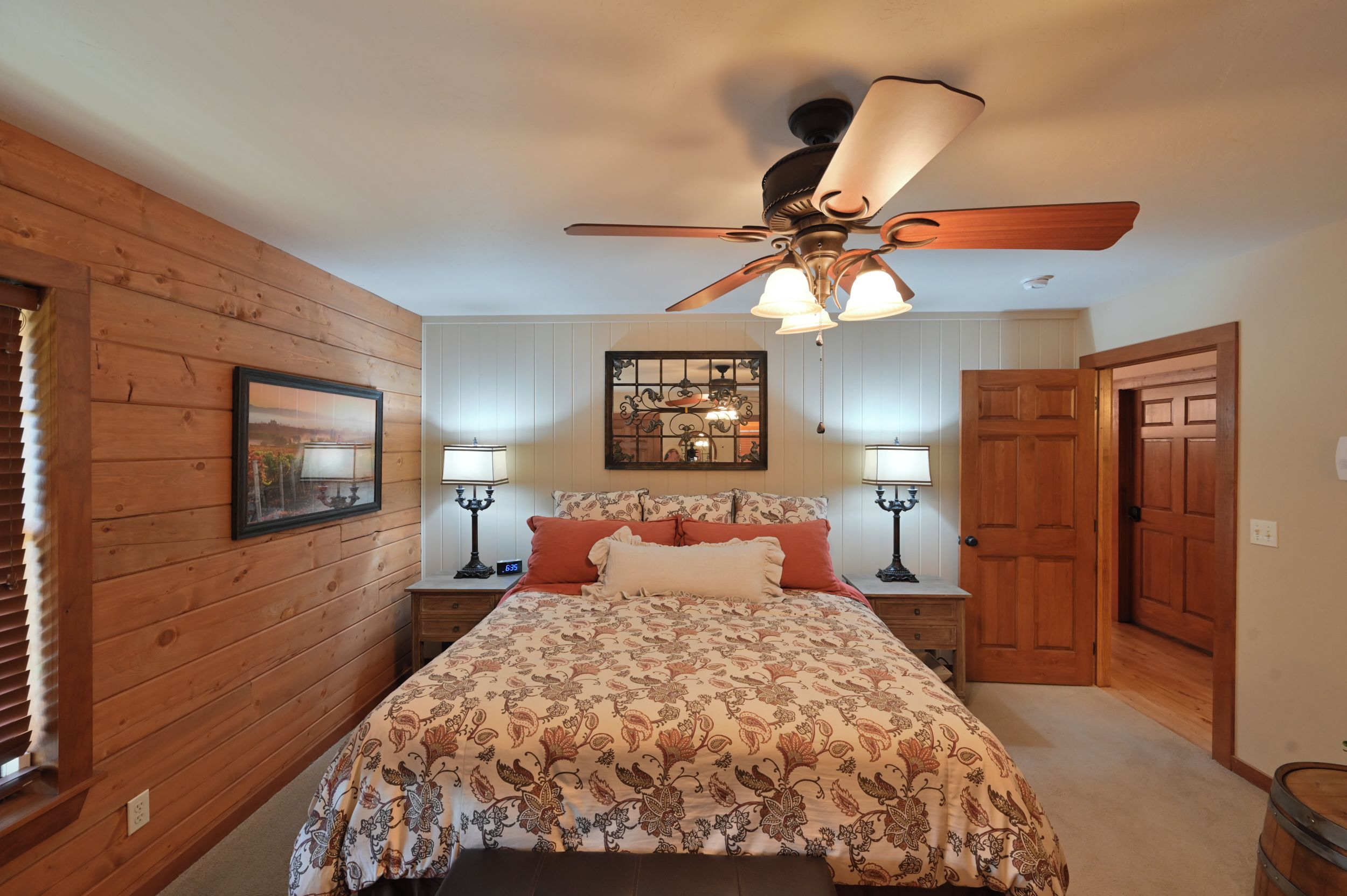 Un dormitorio con ventilador de techo de madera, una cama con ropa de cama floral, dos mesitas de noche con lámparas y una puerta a otra habitación.