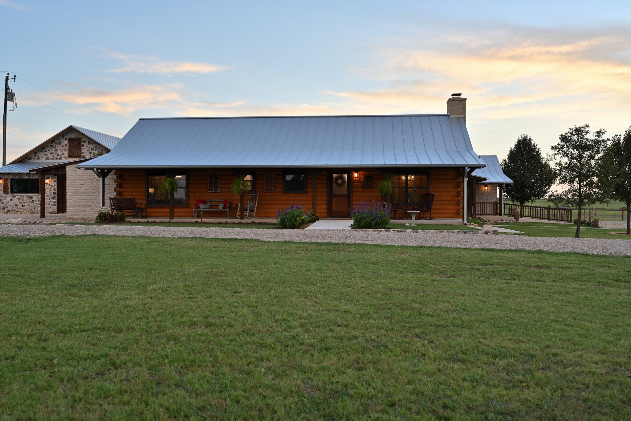 A log cabin with a metal roof, a porch with seating, and a gravel driveway.