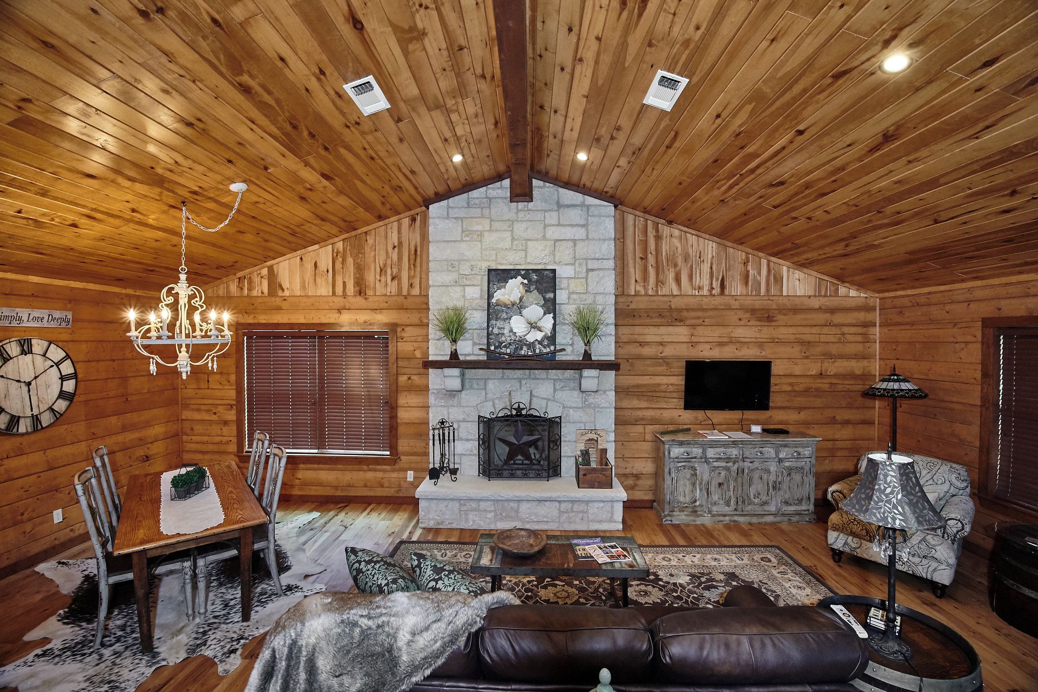 A cozy log cabin interior with a dining area, fireplace, leather sofa, and a chandelier.
