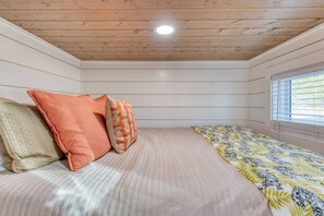 Quarto com cama, almofadas, janela com persianas e teto de madeira.
