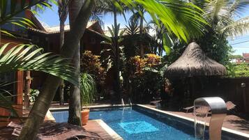 Una piscina con una estructura de techo de paja, rodeada de palmeras y plantas en macetas.