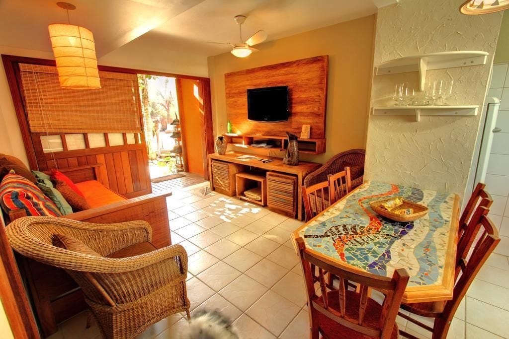 A cozy living area with wicker furniture, a tiled floor, a flat-screen TV mounted on a wooden wall, and a dining table with a mosaic table runner.