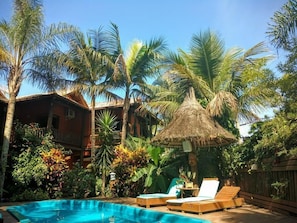 Zona de piscina con sillones de descanso, una estructura de techo de paja y vegetación tropical.