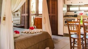 A bedroom with a canopy bed, a dining area with wooden chairs, and a kitchen with white cabinets.
