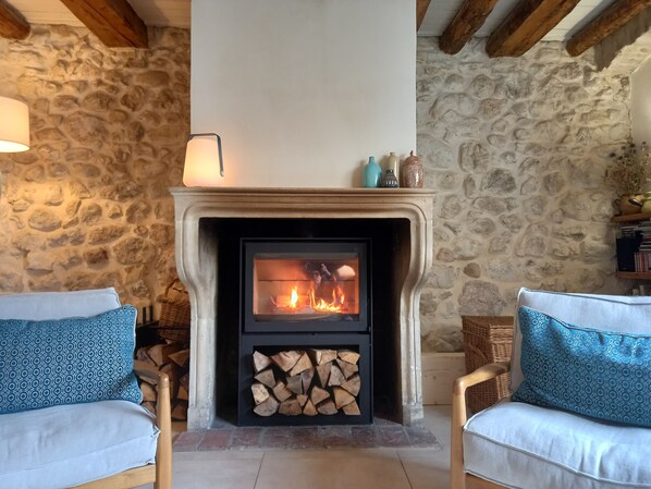 Habitación de paredes de piedra con chimenea, dos sillones con cojines estampados y una pila de leña.