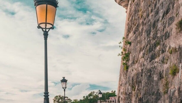 Un muro de piedra con una torre, un sendero pavimentado y un farol.