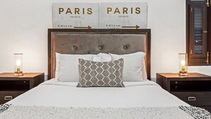 Una cama bien hecha con una colcha blanca, dos almohadas y un cojín decorativo. Hay dos mesitas de noche con lámparas, y un cartel sobre la cama que indica la distancia a París.