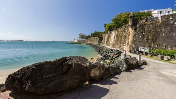 Un paseo marítimo con vistas despejadas al mar, una costa rocosa y un edificio con fachada blanca.
