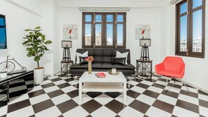 Una sala de estar con piso a cuadros blanco y negro, un sofá negro con cojines blancos, una mesa de centro blanca, una silla roja y dos lámparas de pie.