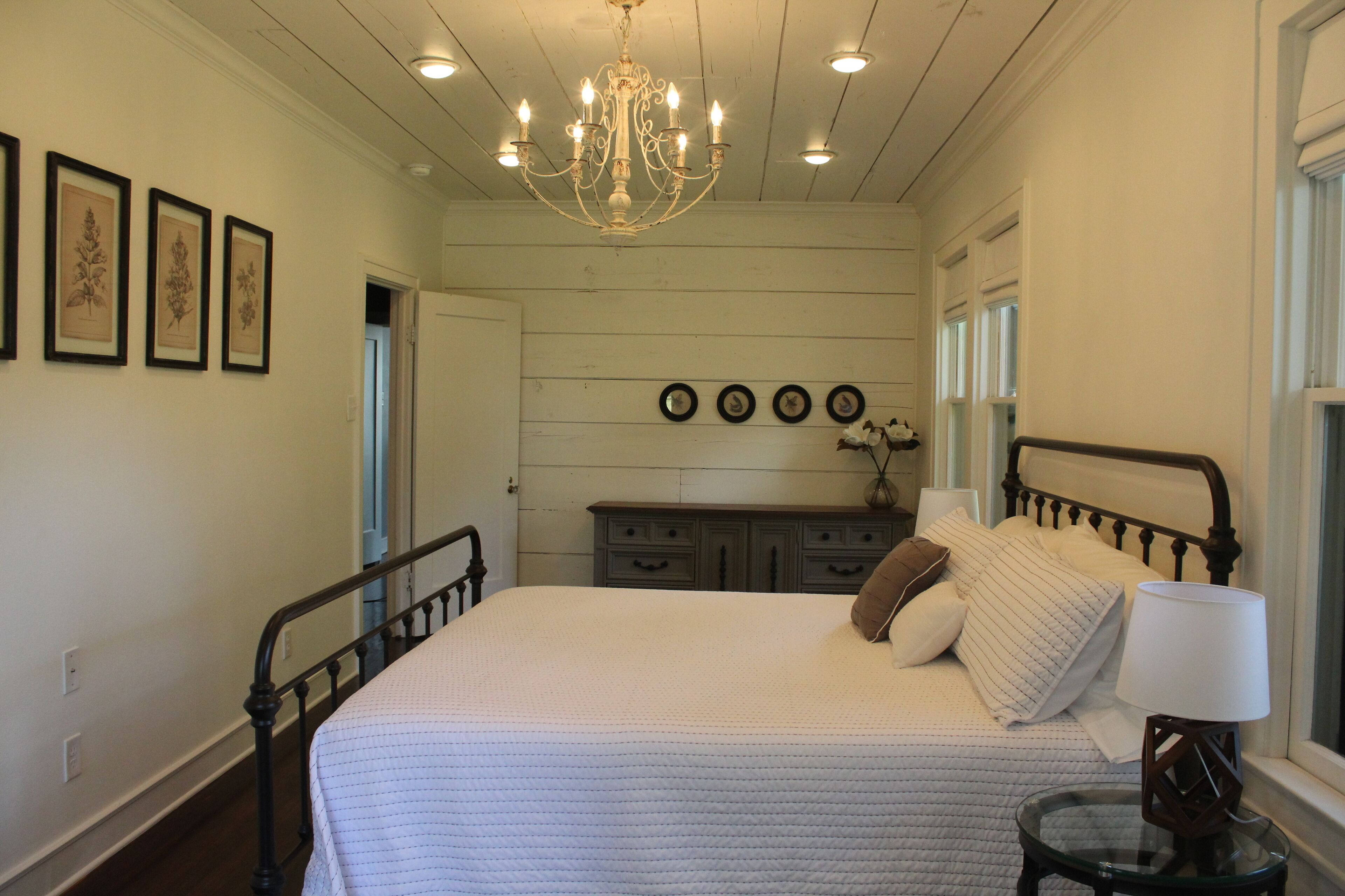 A bedroom with a large bed, a dresser, a chandelier, and framed botanical prints on the wall.