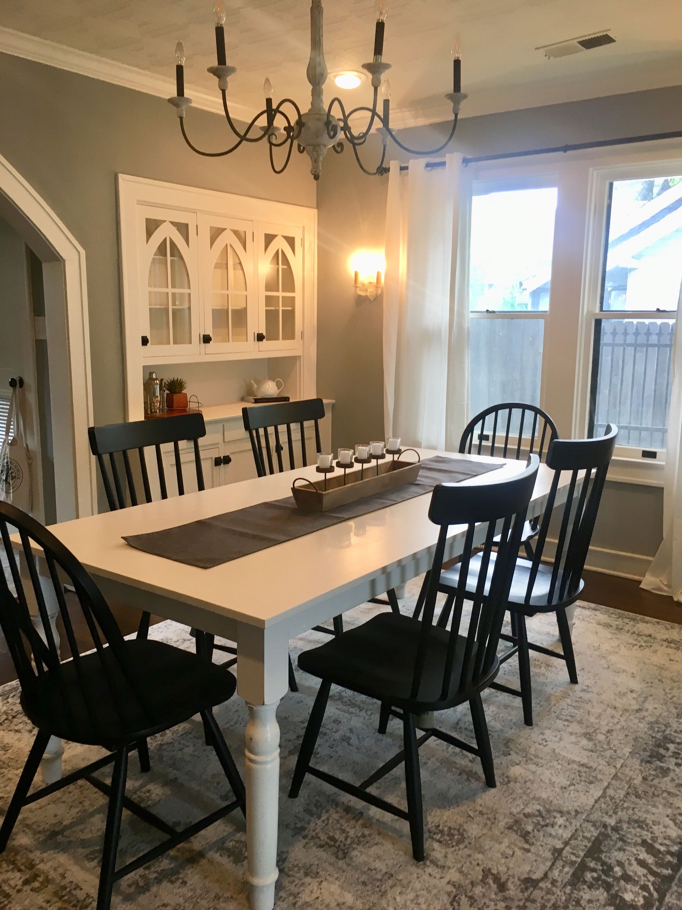 A dining area with a white table, black chairs, a chandelier, and a bay window with curtains.