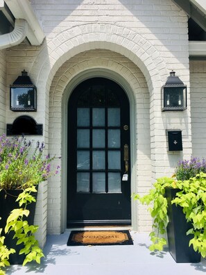 A black arched entrance door with a welcome mat, flanked by two black lantern-style lights and surrounded by potted plants with green and purple foliage. - AS SEEN ON FIXER UPPER "THE BRICK HOUSE" (Waco)