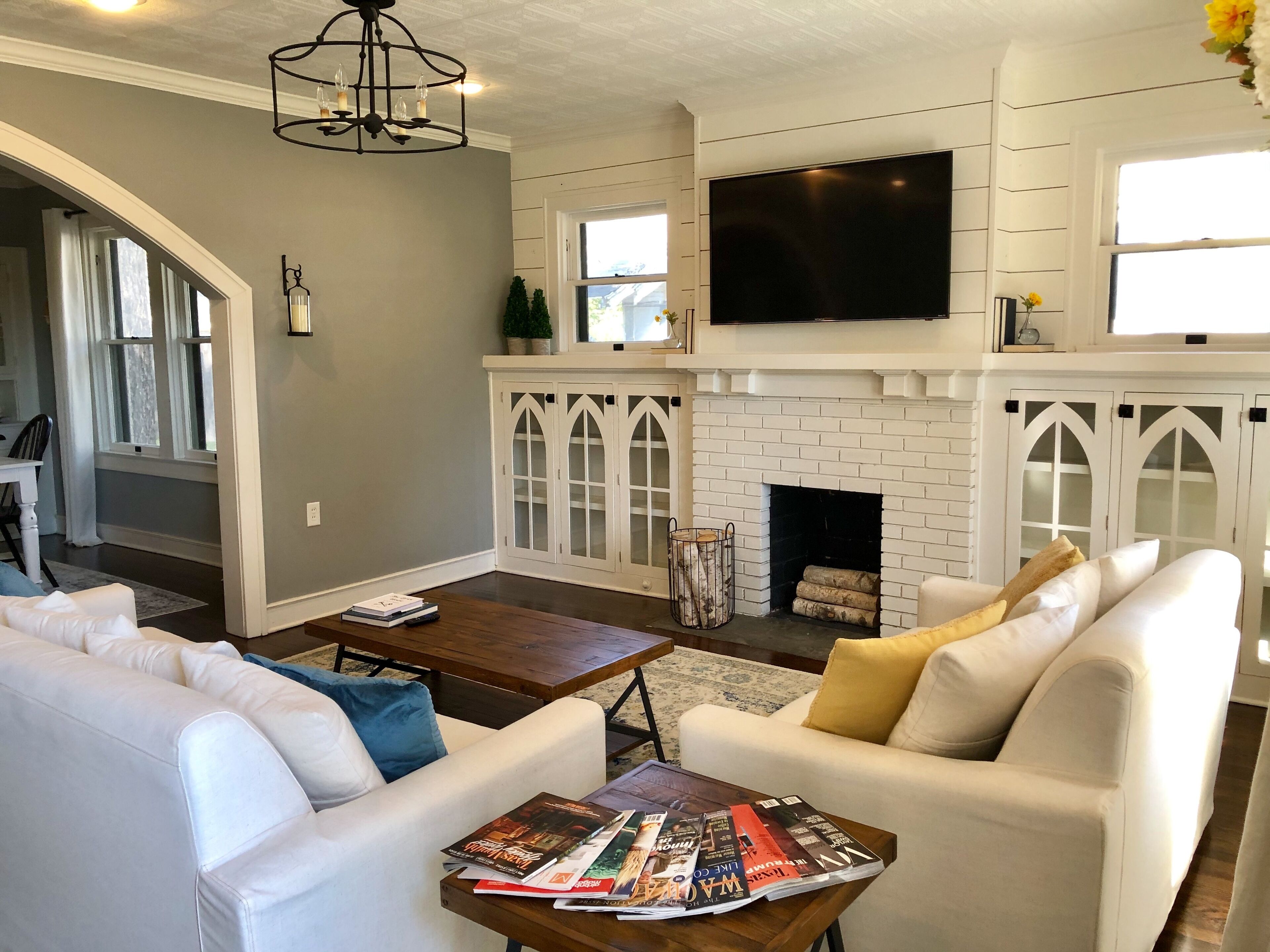 Una sala de estar con chimenea, muebles blancos, televisor de pantalla plana y candelabro.