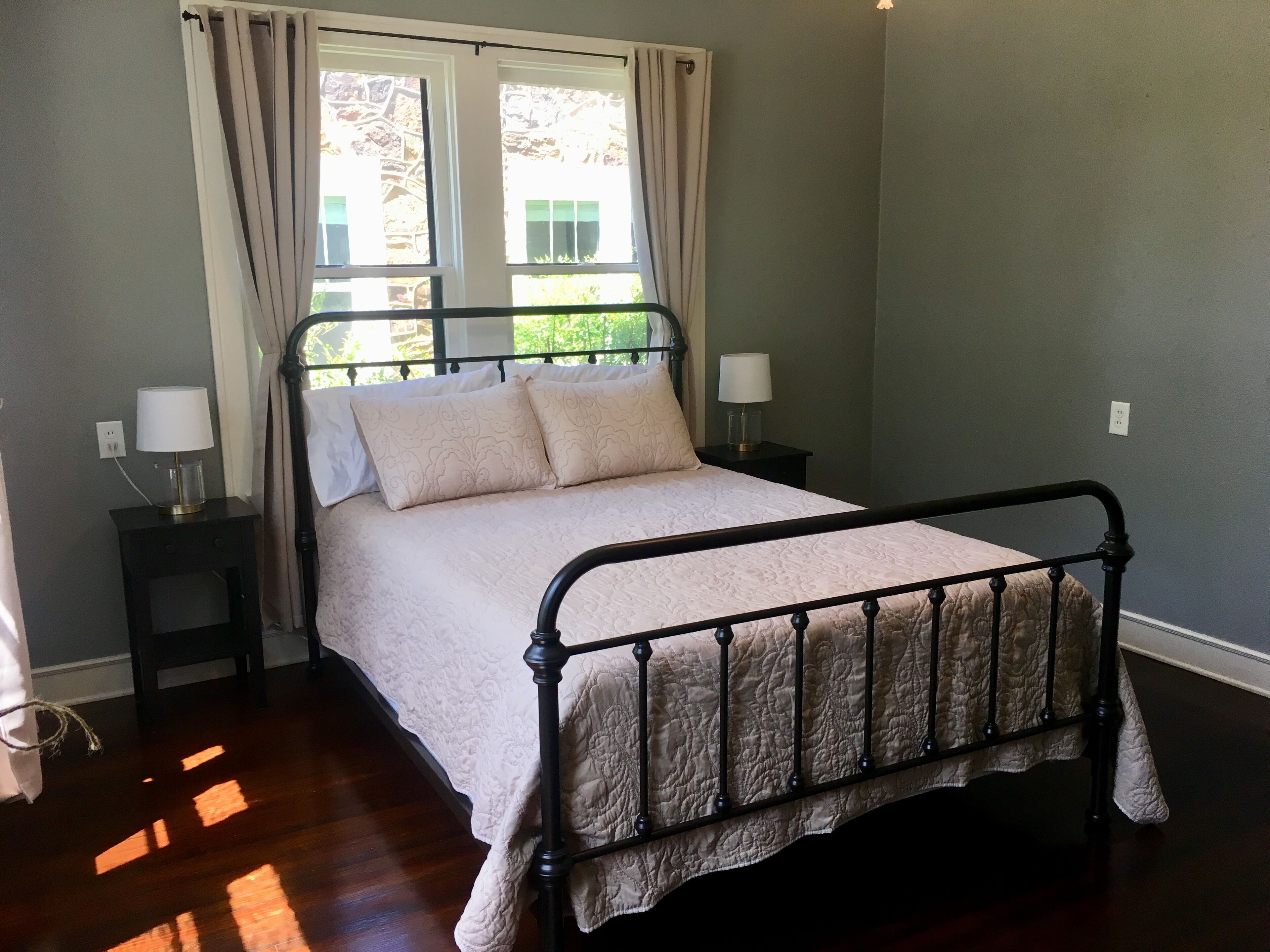 Quarto com cama, duas mesas de cabeceira, janela com cortinas e vista para o verde lá fora.