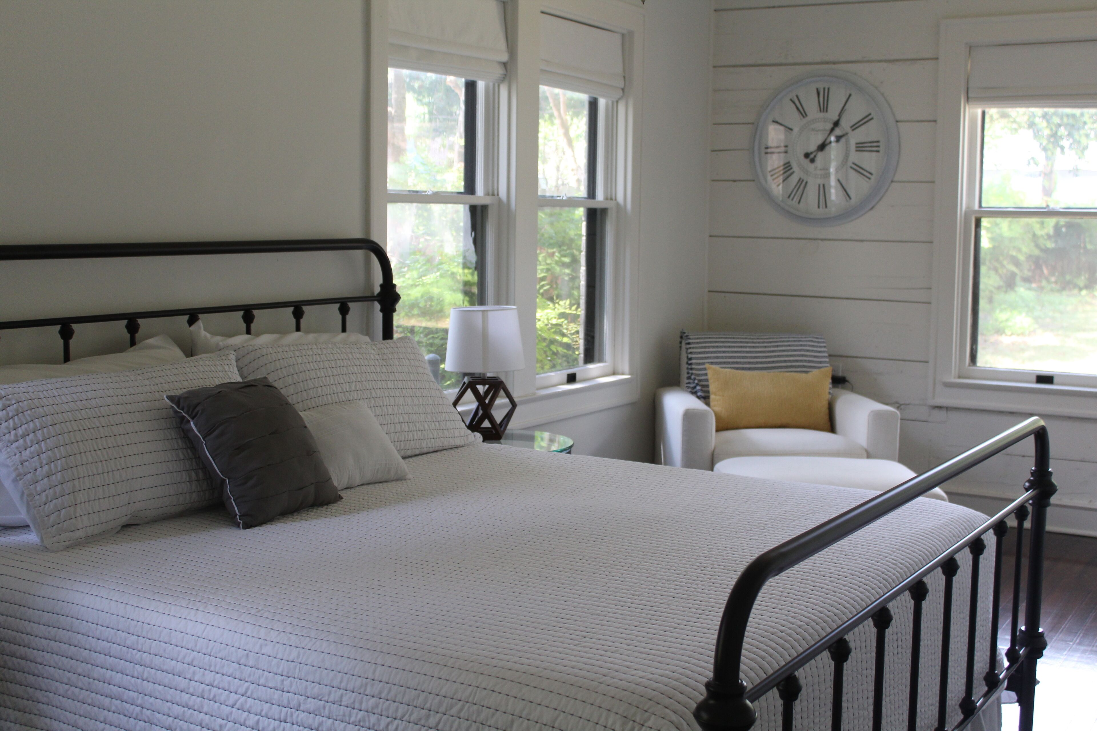 A neatly made bed with a headboard, a bedside lamp, and a chair with cushions.