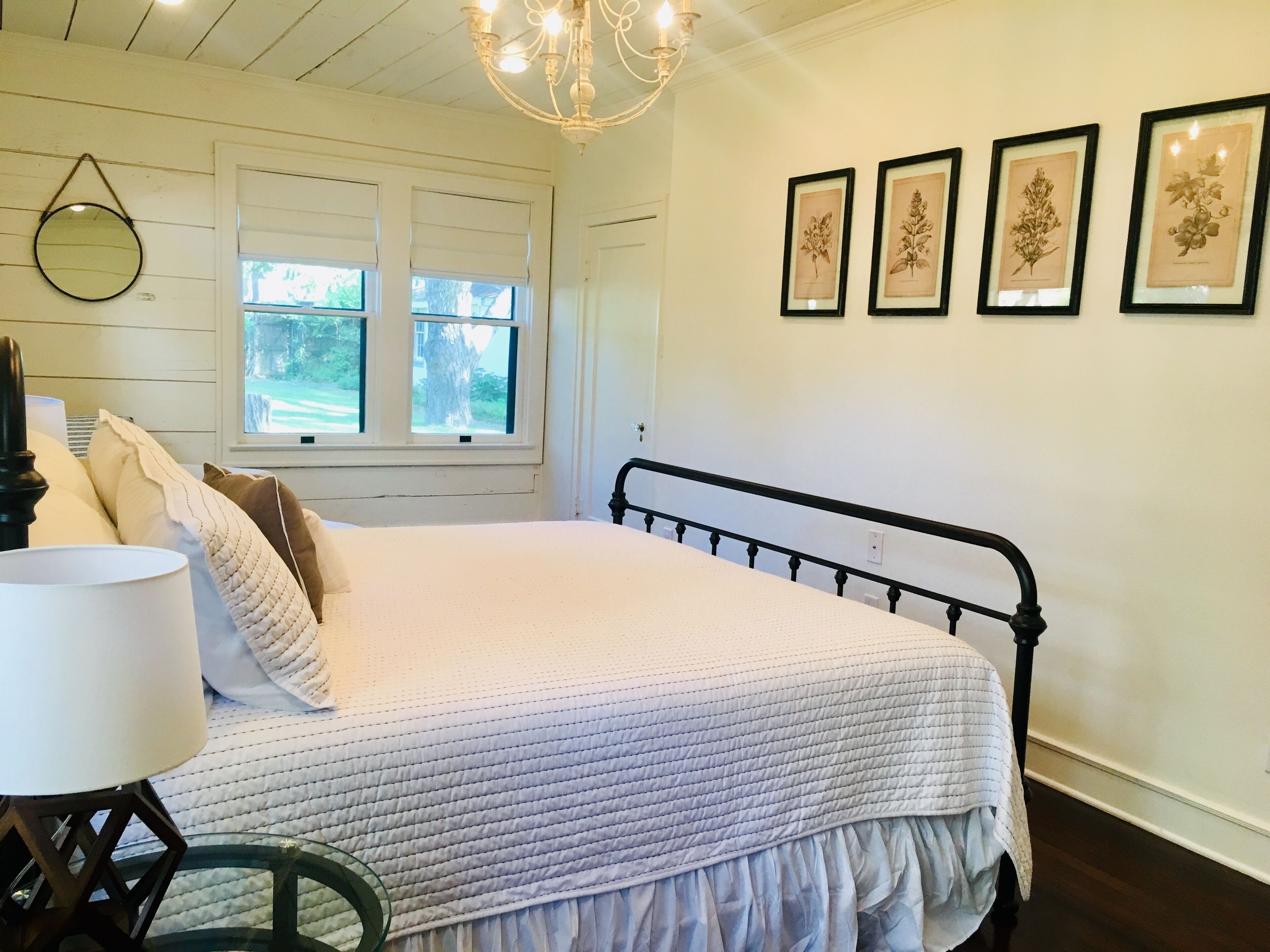 A bedroom with a bed, a round mirror, a chandelier, and framed botanical prints on the wall.