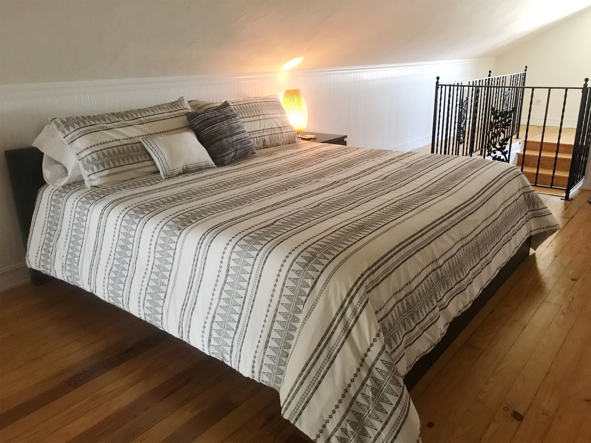 A neatly made bed with patterned bedding, a bedside lamp, and a view of a wooden staircase.