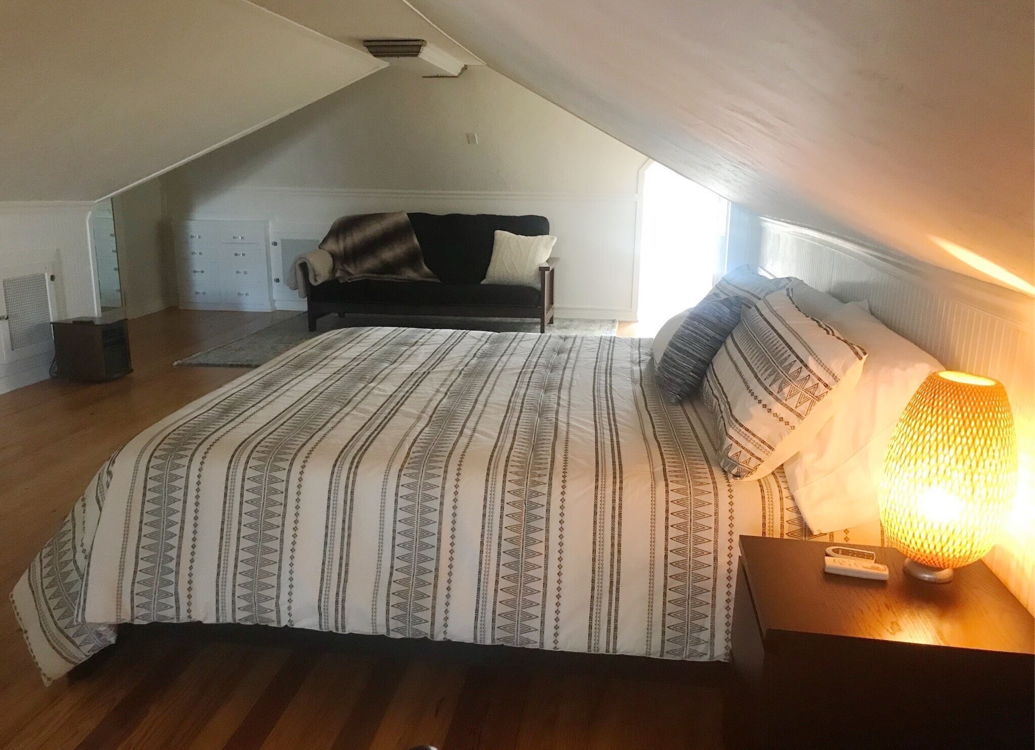 Un dormitorio con techo inclinado, una cama con ropa de cama estampada, una lámpara de noche y un sofá al fondo.