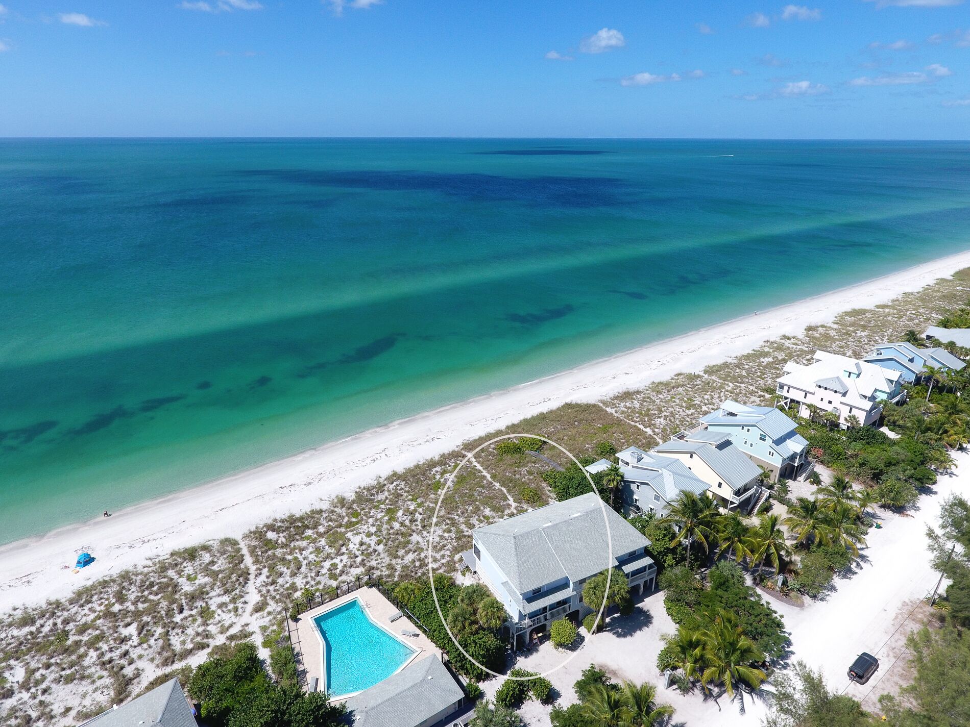 An aerial view of a coastal resort with a clear blue ocean, a sandy beach, and several resort buildings with a swimming pool.