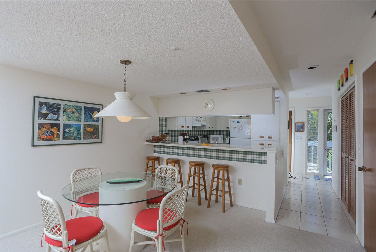 A compact kitchen with white cabinets, a microwave, and a refrigerator. There is a dining area with a glass table and white chairs with red cushions.