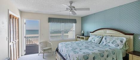 A bedroom with a bed, a nightstand, a chair, a window with blinds, a ceiling fan, and a view of the ocean.