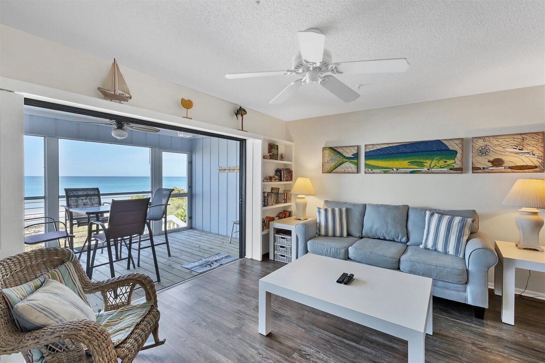 A living room with a view of the ocean, a ceiling fan, a sofa with striped cushions, a coffee table, and a bookshelf.