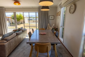 Un comedor con una mesa larga, sillas y un sofá con cojines. Hay cortinas, un reloj y una ventana con vistas al exterior.