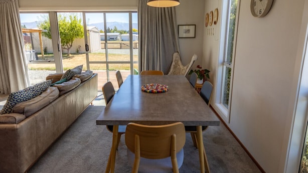 A dining area with a long table, chairs, and a sofa with cushions. There are curtains, a clock, and a window with a view of the outside. - Rejuvenate on Rata · Rejuvenate on Rata, Twizel FREE WIFI (Twizel)