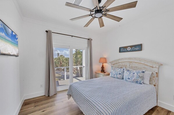 A bedroom with a bed, a ceiling fan, a nightstand with a lamp, a beach-themed sign, and a view of the outdoors through a sliding door.