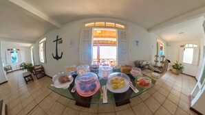 A dining area with a glass table surrounded by various food containers, a potted plant, and a view of the outside through a window. - Pousada Marinheiro (Cairu)